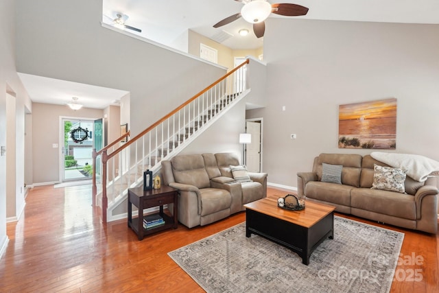 living room with stairs, a towering ceiling, wood finished floors, baseboards, and a ceiling fan