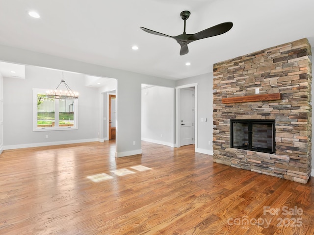 unfurnished living room with baseboards, light wood-style flooring, ceiling fan with notable chandelier, and a stone fireplace