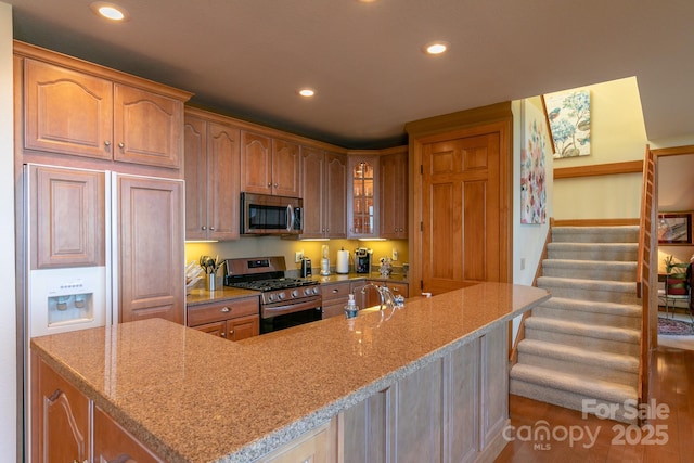kitchen featuring appliances with stainless steel finishes, recessed lighting, light stone counters, glass insert cabinets, and an island with sink