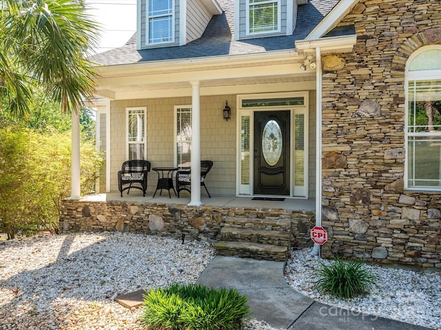 view of exterior entry with covered porch, roof with shingles, and stone siding