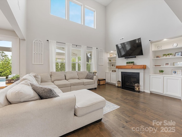 living area featuring dark wood-type flooring, a fireplace with flush hearth, a towering ceiling, and a healthy amount of sunlight