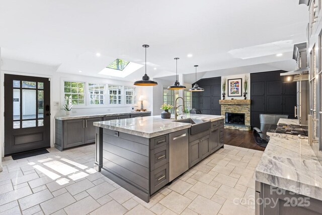kitchen with stainless steel dishwasher, a stone fireplace, a large island with sink, a sink, and a skylight