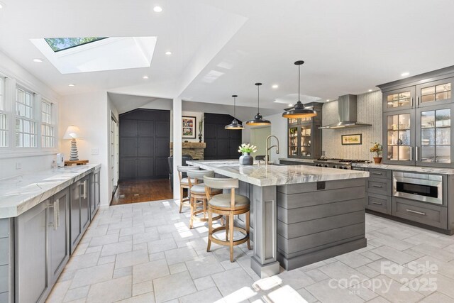 kitchen featuring light stone countertops, glass insert cabinets, wall chimney range hood, gray cabinets, and a skylight