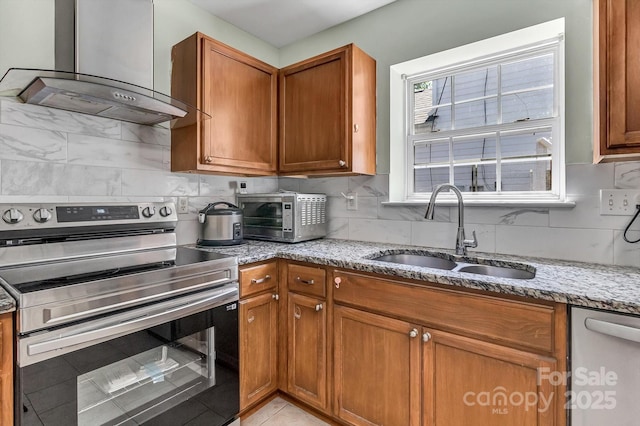 kitchen with appliances with stainless steel finishes, a sink, wall chimney exhaust hood, brown cabinets, and light stone counters
