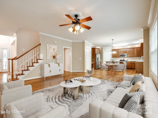 living area with crown molding, light wood-style flooring, ceiling fan with notable chandelier, baseboards, and stairway