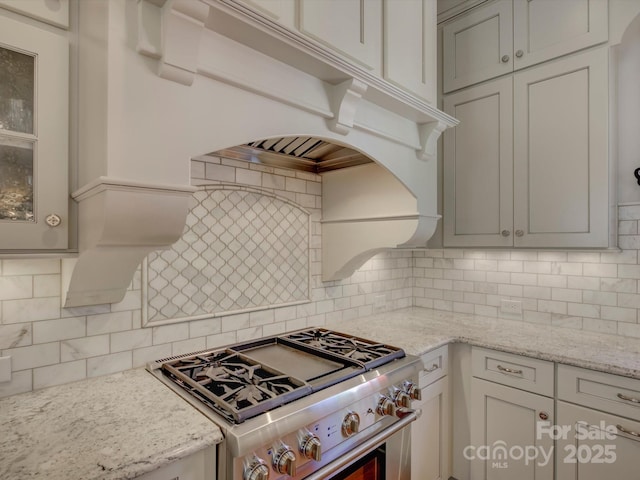 kitchen featuring high end stove, backsplash, light stone countertops, and glass insert cabinets