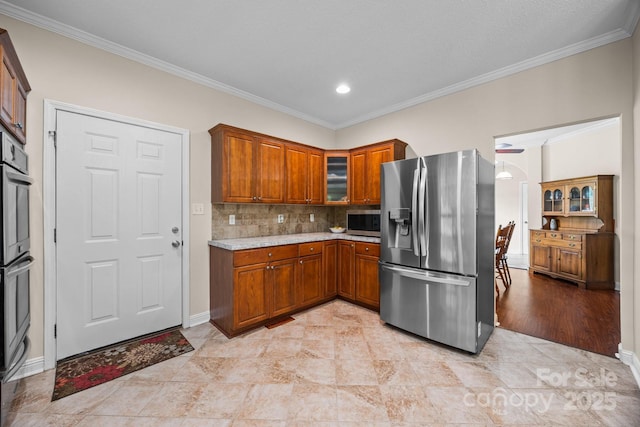 kitchen with appliances with stainless steel finishes, ornamental molding, brown cabinetry, glass insert cabinets, and decorative backsplash