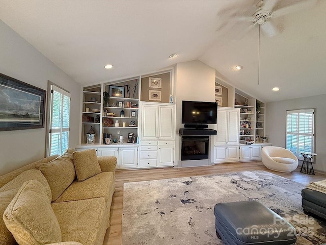 living area featuring ceiling fan, light wood finished floors, a fireplace, built in shelves, and vaulted ceiling