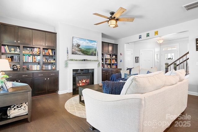living area with a ceiling fan, dark wood-style flooring, a glass covered fireplace, baseboards, and stairs