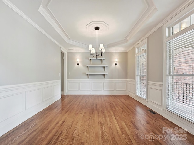 unfurnished dining area with visible vents, wood finished floors, a raised ceiling, a wainscoted wall, and an inviting chandelier