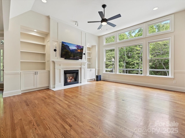 unfurnished living room with a glass covered fireplace, baseboards, light wood-type flooring, built in shelves, and a ceiling fan