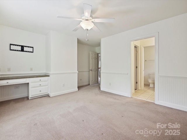 unfurnished bedroom featuring light colored carpet, built in study area, a ceiling fan, baseboards, and ensuite bathroom
