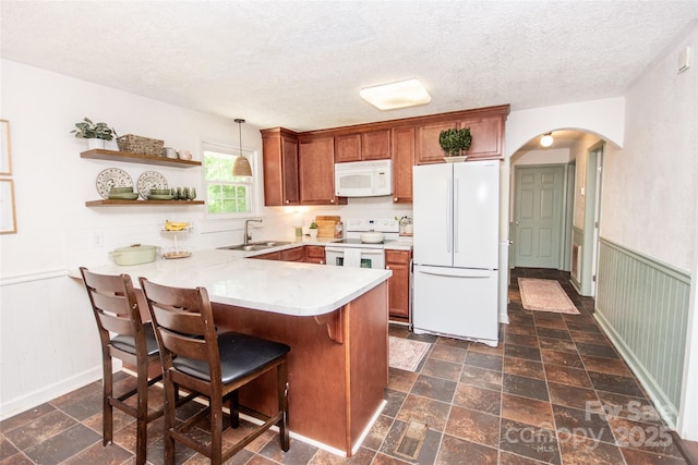 kitchen with brown cabinetry, a peninsula, white appliances, arched walkways, and a sink
