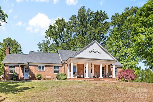 greek revival inspired property with covered porch, a chimney, a front yard, and brick siding