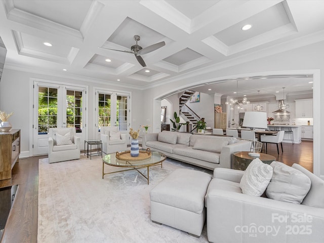 living area featuring arched walkways, ceiling fan with notable chandelier, stairs, wood finished floors, and coffered ceiling
