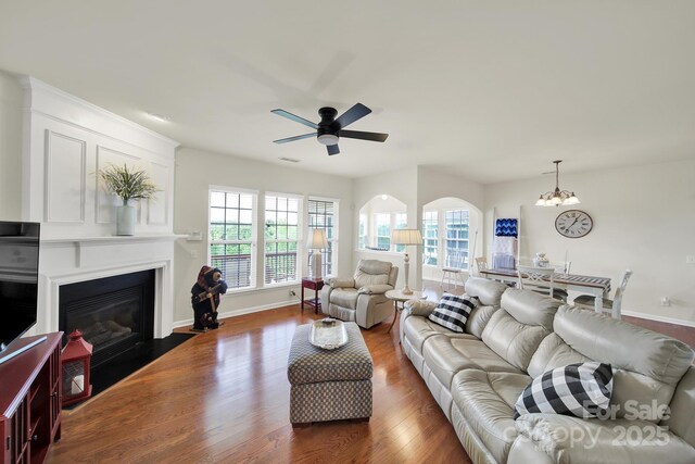living room with baseboards, a fireplace with flush hearth, ceiling fan with notable chandelier, and dark wood-style floors