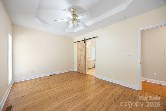 unfurnished bedroom featuring a raised ceiling, light wood-style floors, a barn door, ensuite bath, and baseboards