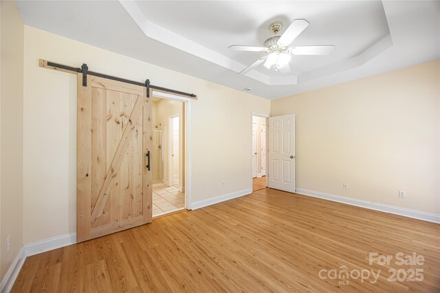 empty room with a raised ceiling, light wood finished floors, a barn door, a ceiling fan, and baseboards