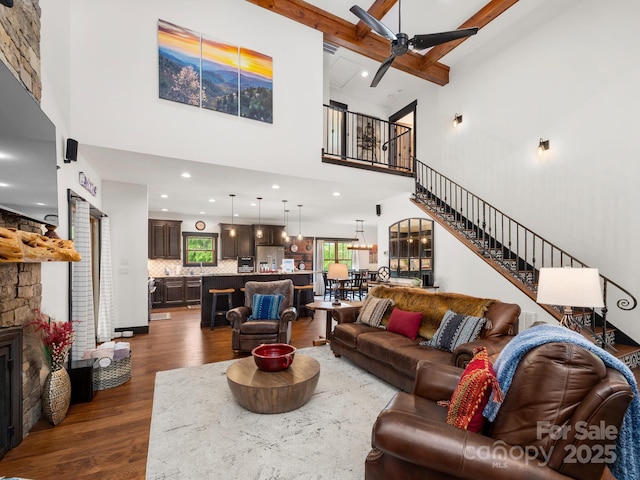 living area with dark wood-type flooring, a high ceiling, stairs, a ceiling fan, and beamed ceiling