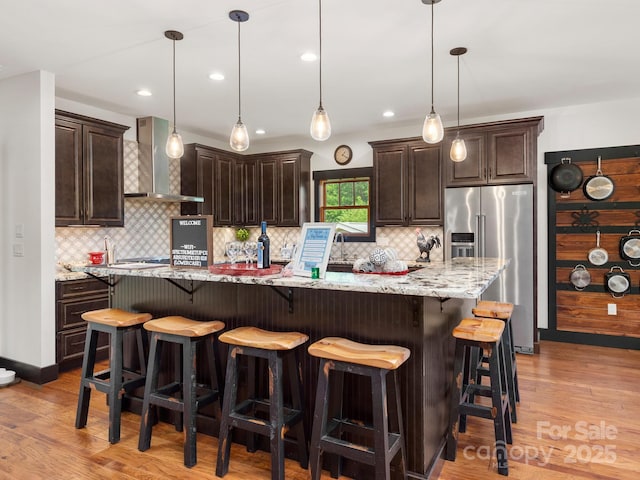 kitchen with high end fridge, wall chimney exhaust hood, dark brown cabinets, light wood-style floors, and recessed lighting
