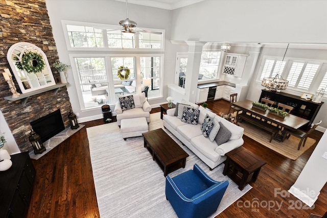 living area with dark wood-style flooring, a stone fireplace, ceiling fan with notable chandelier, baseboards, and a towering ceiling