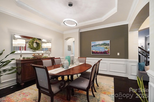dining area with wood finished floors, a tray ceiling, wainscoting, arched walkways, and ornamental molding