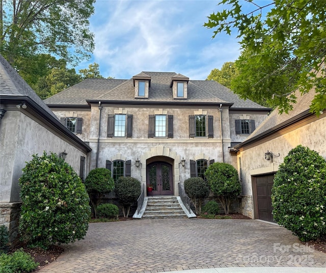 french country home with decorative driveway, roof with shingles, stucco siding, french doors, and an attached garage