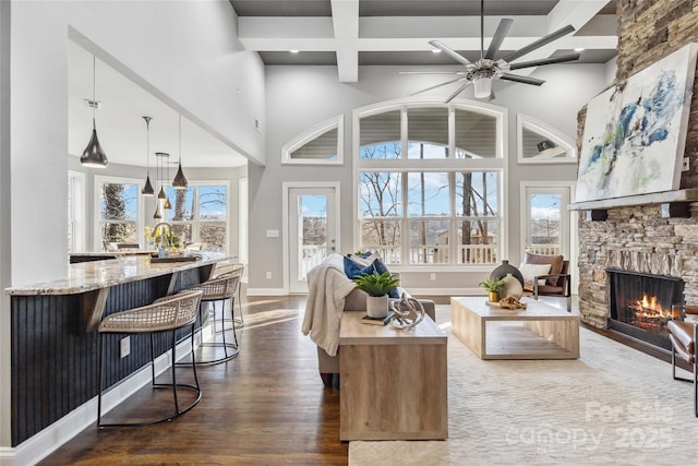 living room featuring ceiling fan, a stone fireplace, a towering ceiling, dark wood finished floors, and beamed ceiling