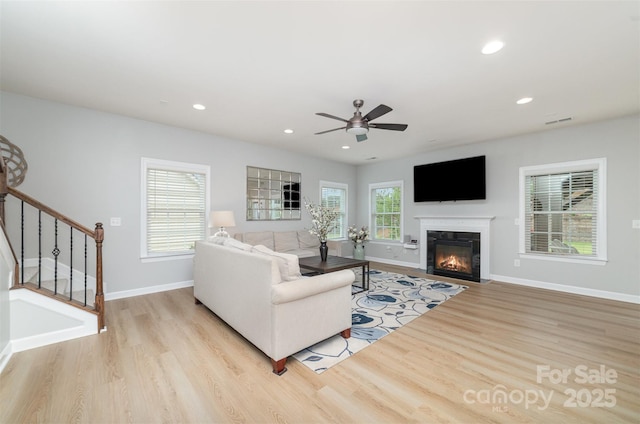 living area featuring a ceiling fan, baseboards, a glass covered fireplace, light wood-style floors, and stairs