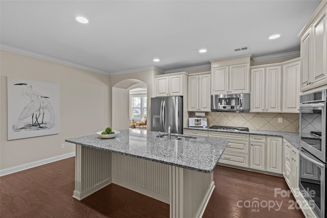kitchen featuring light stone counters, a sink, arched walkways, stainless steel appliances, and a center island with sink