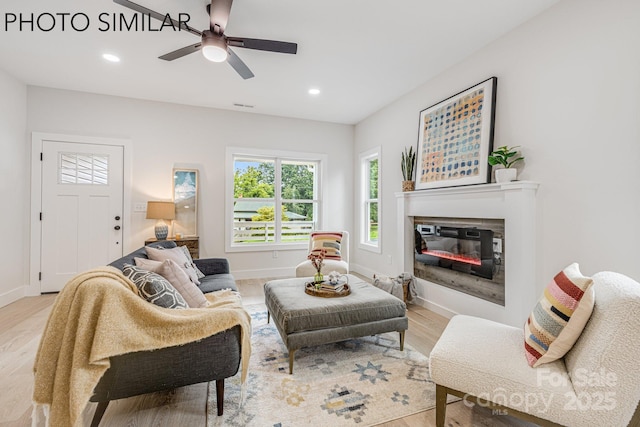living room with visible vents, light wood finished floors, a ceiling fan, baseboards, and a glass covered fireplace