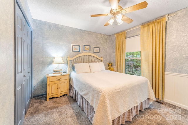 carpeted bedroom with a closet, a textured ceiling, a ceiling fan, wainscoting, and wallpapered walls