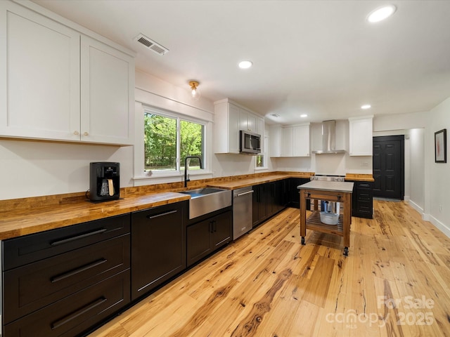 kitchen featuring wall chimney range hood, white cabinets, a sink, wooden counters, and stainless steel appliances