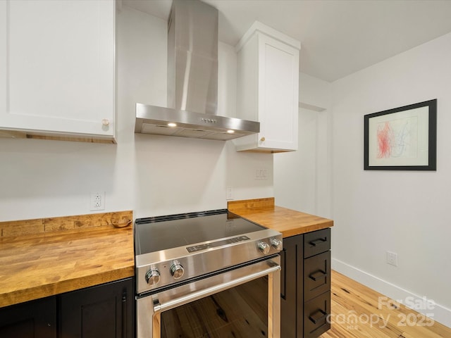 kitchen with butcher block countertops, wall chimney range hood, stainless steel electric range, white cabinets, and light wood finished floors