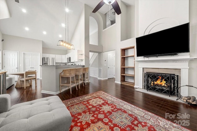 living area with high vaulted ceiling, dark wood-style flooring, built in shelves, ceiling fan, and a lit fireplace