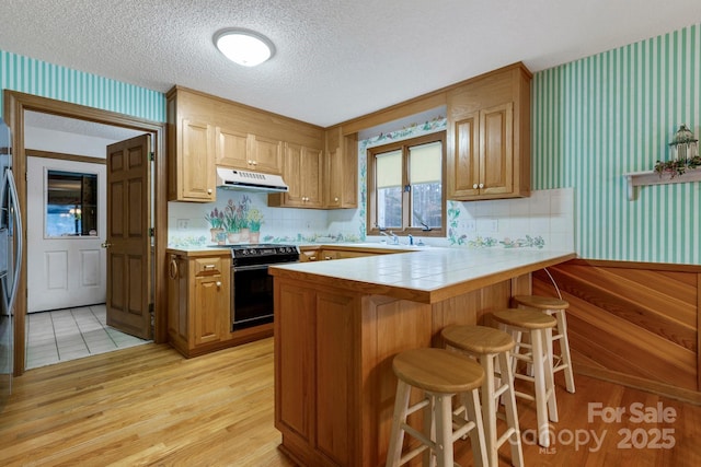 kitchen with a peninsula, a kitchen breakfast bar, a textured ceiling, under cabinet range hood, and black electric range