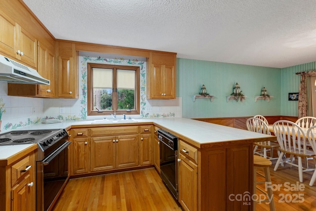 kitchen with under cabinet range hood, a sink, a peninsula, black appliances, and light wood-style floors