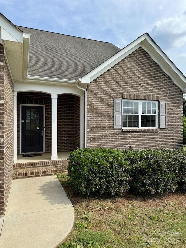 doorway to property featuring brick siding, roof with shingles, and a porch