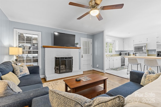living room featuring crown molding, light wood-style flooring, a brick fireplace, a ceiling fan, and baseboards