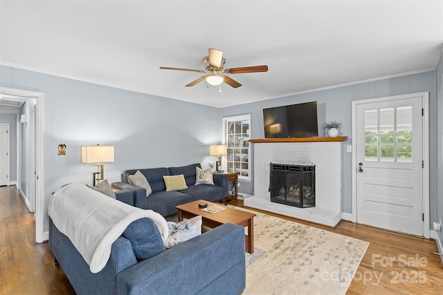 living area featuring wood finished floors, ceiling fan, ornamental molding, and a brick fireplace