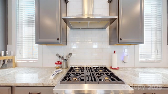 kitchen featuring wall chimney range hood, light stone countertops, stovetop with downdraft, decorative backsplash, and gray cabinetry