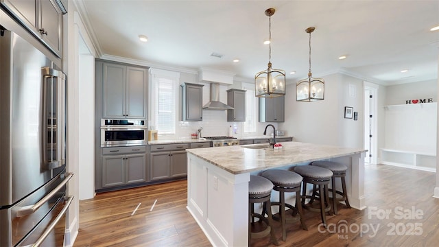 kitchen with gray cabinetry, appliances with stainless steel finishes, a sink, and wall chimney exhaust hood