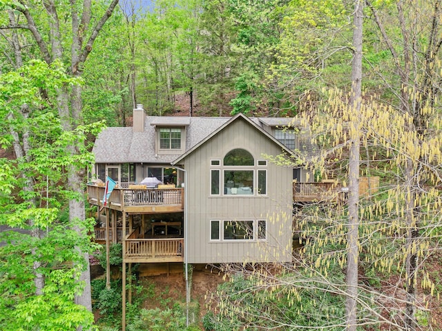 back of property with a view of trees, roof with shingles, a wooden deck, and a chimney