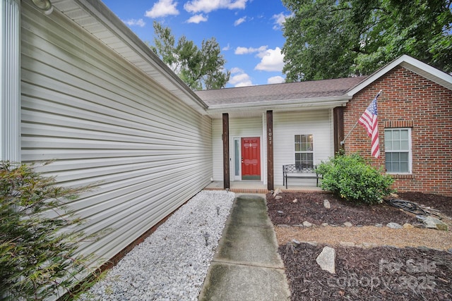property entrance featuring brick siding, roof with shingles, and covered porch