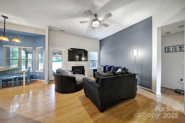 living area with ceiling fan, light wood-style floors, a tiled fireplace, baseboards, and a textured ceiling