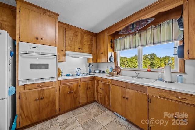 kitchen featuring brown cabinetry, white appliances, light countertops, under cabinet range hood, and a sink