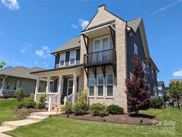 traditional-style house with ceiling fan, a balcony, covered porch, a front yard, and a chimney