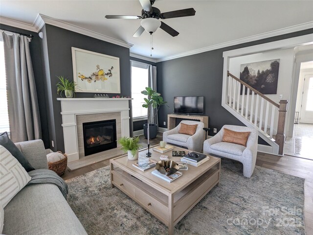 living room featuring crown molding, a glass covered fireplace, wood finished floors, ceiling fan, and stairway