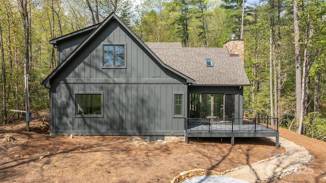 exterior space featuring roof with shingles, a wooden deck, board and batten siding, a view of trees, and a chimney