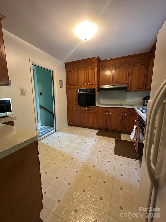 kitchen featuring black oven, brown cabinetry, freestanding refrigerator, stainless steel microwave, and under cabinet range hood